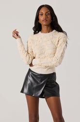 A woman is wearing a cream-colored, textured knit sweater with pearl embellishments throughout. The sweater has a round neckline and long sleeves. She is also wearing a black faux leather mini skirt with an asymmetrical design and a small buckle detail on the waistband.